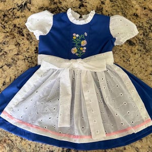 Puede incluir: Un vestido azul con mangas cortas de encaje blanco y un delantal de encaje blanco a juego con un gran lazo atado en la cintura. El corpi&ntilde;o muestra un bordado floral central con flores rosas, amarillas y blancas, y el dobladillo tiene un borde de cinta rosa.