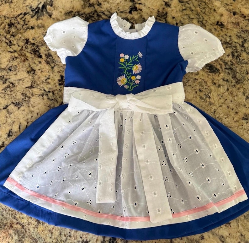 Puede incluir: Un vestido azul con mangas cortas de encaje blanco y un delantal de encaje blanco a juego con un gran lazo atado en la cintura. El corpi&ntilde;o muestra un bordado floral central con flores rosas, amarillas y blancas, y el dobladillo tiene un borde de cinta rosa.