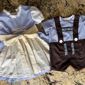 Puede incluir: Un vestido de cuadros vichy azul y blanco con mangas abullonadas de encaje blanco y un delantal color crema, junto a una camisa de cuadros vichy azul y blanco con pantalones cortos marrones con tirantes, botones blancos y bordados florales.