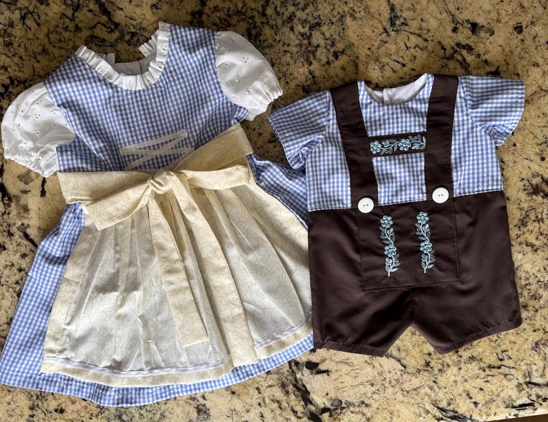 Puede incluir: Un vestido de cuadros vichy azul y blanco con mangas abullonadas de encaje blanco y un delantal color crema, junto a una camisa de cuadros vichy azul y blanco con pantalones cortos marrones con tirantes, botones blancos y bordados florales.