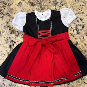 Pode incluir: Um vestido preto e vermelho com mangas de ilh&oacute;s brancas e uma gola com babados branca. O vestido apresenta um la&ccedil;o vermelho na cintura e uma saia vermelha com acabamento preto e branco. O corpete tem um design decorativo cruzado.