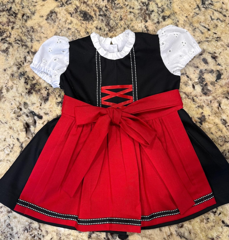 Pode incluir: Um vestido preto e vermelho com mangas de ilh&oacute;s brancas e uma gola com babados branca. O vestido apresenta um la&ccedil;o vermelho na cintura e uma saia vermelha com acabamento preto e branco. O corpete tem um design decorativo cruzado.