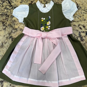 Pode incluir: Vestido verde-oliva com mangas de ilh&oacute;s brancas e gola franzida branca. O corpete apresenta bordados florais. Um avental e um la&ccedil;o rosa completam o visual tradicional. O vestido &eacute; projetado para uma crian&ccedil;a.