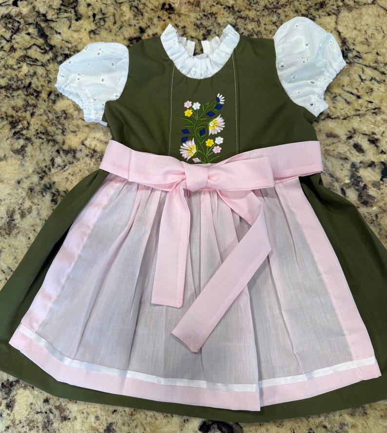 Pode incluir: Vestido verde-oliva com mangas de ilh&oacute;s brancas e gola franzida branca. O corpete apresenta bordados florais. Um avental e um la&ccedil;o rosa completam o visual tradicional. O vestido &eacute; projetado para uma crian&ccedil;a.