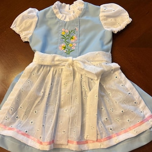 Pode incluir: Um vestido azul claro com mangas brancas de renda e um avental branco. O corpete apresenta um design floral bordado. Uma fita rosa adorna a bainha do avental. O vestido tem um la&ccedil;o branco na cintura.