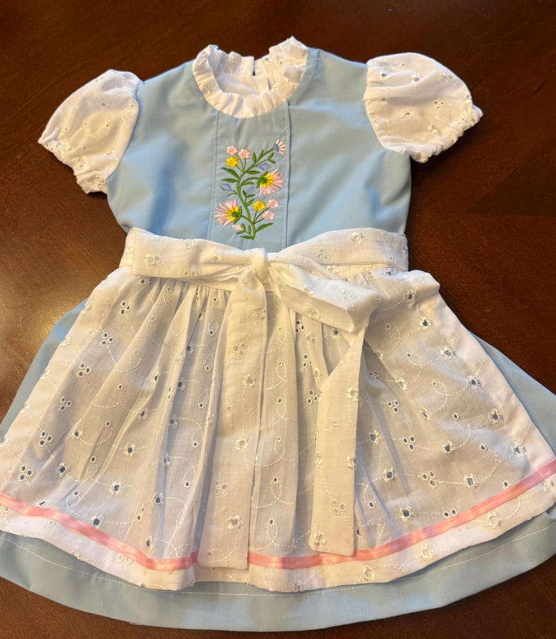 Pode incluir: Um vestido azul claro com mangas brancas de renda e um avental branco. O corpete apresenta um design floral bordado. Uma fita rosa adorna a bainha do avental. O vestido tem um la&ccedil;o branco na cintura.