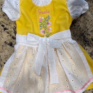 Puede incluir: Un vestido amarillo brillante con mangas abullonadas de encaje blanco y un delantal blanco a juego con lazo en la cintura. El corpi&ntilde;o muestra el nombre Susie bordado en rosa sobre un dise&ntilde;o floral colorido con flores rosas y azules.