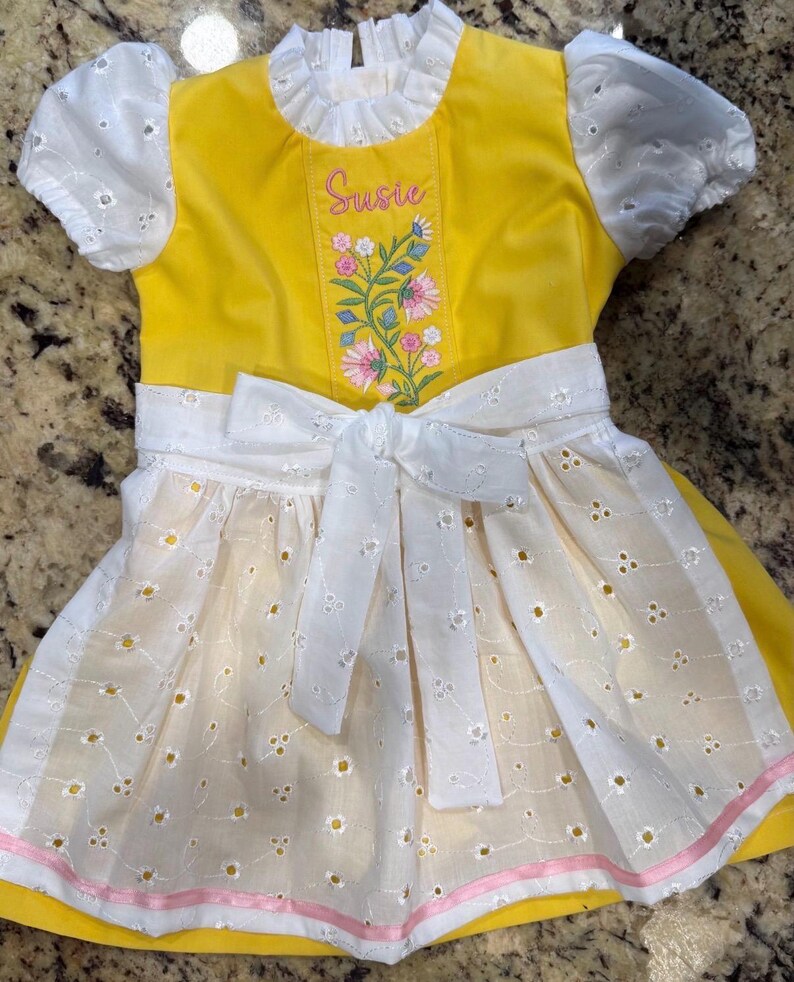 Puede incluir: Un vestido amarillo brillante con mangas abullonadas de encaje blanco y un delantal blanco a juego con lazo en la cintura. El corpi&ntilde;o muestra el nombre Susie bordado en rosa sobre un dise&ntilde;o floral colorido con flores rosas y azules.