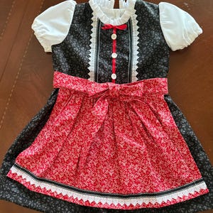 Pode incluir: Vestido de estilo tradicional com corpete preto e saia floral vermelha. O vestido apresenta mangas bufantes brancas, gola de renda branca e la&ccedil;o vermelho. A saia tem um padr&atilde;o floral vermelho com acabamento em renda branca. Para crian&ccedil;a.