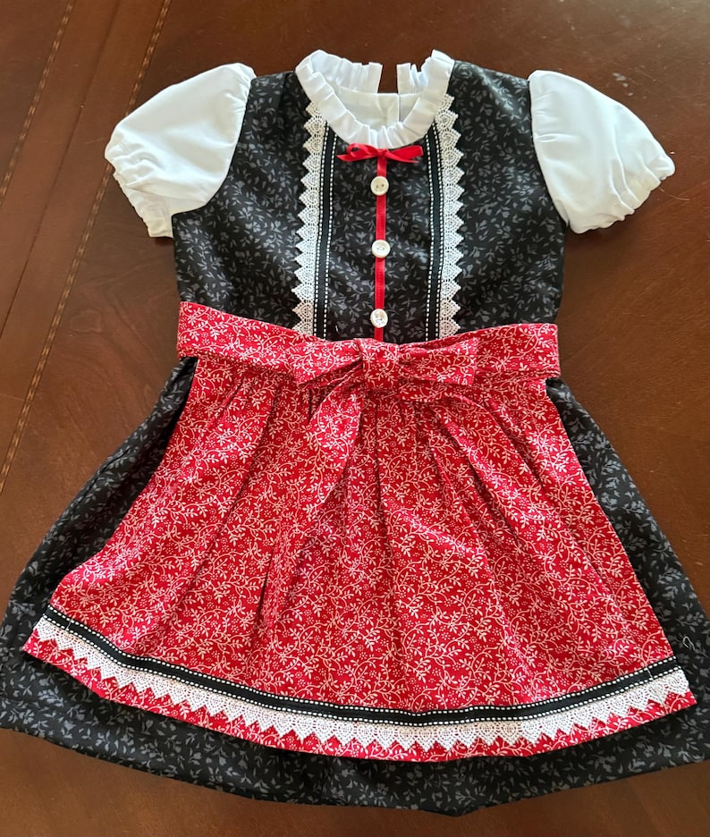 Pode incluir: Vestido de estilo tradicional com corpete preto e saia floral vermelha. O vestido apresenta mangas bufantes brancas, gola de renda branca e la&ccedil;o vermelho. A saia tem um padr&atilde;o floral vermelho com acabamento em renda branca. Para crian&ccedil;a.