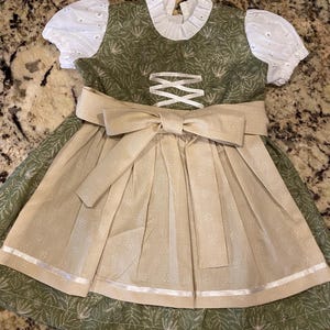 Pode incluir: Um vestido dirndl verde e bege com um grande la&ccedil;o. O vestido apresenta mangas de ilh&oacute;s brancas e uma gola com babados brancos. O corpete tem um design cruzado e a saia tem um avental bege com detalhes florais brancos.