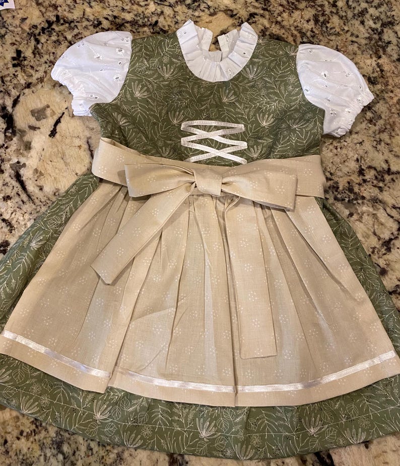 Pode incluir: Um vestido dirndl verde e bege com um grande la&ccedil;o. O vestido apresenta mangas de ilh&oacute;s brancas e uma gola com babados brancos. O corpete tem um design cruzado e a saia tem um avental bege com detalhes florais brancos.