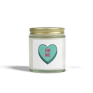 May include: A white candle jar with a gold lid. The label on the jar features a green heart with the text "DM ME" in pink.