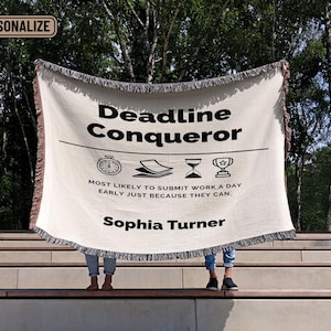 Może przedstawiać: Biały koc z czarnym tekstem, który brzmi "Deadline Conqueror" i "Most likely to submit work a day early just because they can." Tekst zawiera również stoper, stos papierów, klepsydrę i puchar. Imię "Sophia Turner" jest nadrukowane na dole koca.