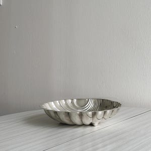 Könnte beinhalten: Eine silberfarbene, muschelförmige Schale mit geriffeltem Design. Die Schale hat eine abgerundete Form und steht auf drei kleinen Füßen. Das Innere der Schale hat eine reflektierende Oberfläche. Die Schale ist wahrscheinlich aus Metall.