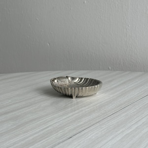 Könnte beinhalten: Eine kleine, silberfarbene Metallschale in Form einer Muschel. Die Schale weist eine gerippte, fächerartige Textur auf ihrer Oberfläche auf und wird von drei kurzen, integrierten Füßen getragen, die den Boden leicht anheben.