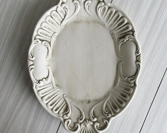 May include: An oval, off-white decorative tray with ornate, raised scrollwork and shell-like details around the rim. The tray has a slightly aged appearance, with subtle shading and a vintage aesthetic. The design is symmetrical and elegant.