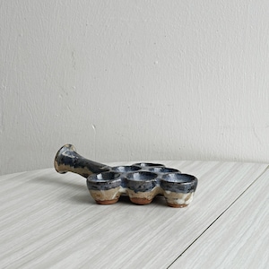 May include: Handmade ceramic tasting flight set. The set features a handle and six small cups. The cups and handle are glazed in shades of blue and brown. The set is designed for serving small portions of beverages.