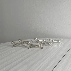 May include: A collection of six silver-colored, stylized cheetah figurines. The cheetahs are in a running pose, with their legs extended. The figurines are placed on a light-colored, textured surface, with a gray wall in the background.