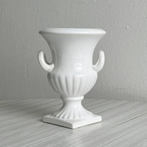 以下が含まれることがあります： 光沢のある白い陶器製の壺型花瓶。広がった縁、2つの湾曲したサイドハンドル、リブ状のボディデザインが特徴です。溝のあるテクスチャーが施された正方形の台座の上に立ち、全体的に清潔感のある無地の白い仕上げとなっています。
