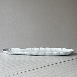 May include: A long, white ceramic serving platter with a scalloped edge and a subtle, embossed leaf-like pattern across its surface. The dish has an elongated, oval shape with a glossy finish and a slightly raised rim, presenting a clean, neutral look.