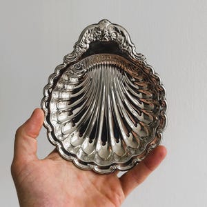 Può includere: Un piatto d'argento a forma di conchiglia con un design decorativo e ornato attorno al bordo. Il piatto è tenuto nella mano di una persona.
