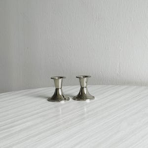 Op de afbeelding: Twee zilverkleurige kandelaars met een klassiek ontwerp. Elke houder heeft een brede, ronde basis, een slanke steel en een kleine, cirkelvormige bovenkant om een kaars in te plaatsen. De houders staan op een lichtgekleurd oppervlak, wat duidt op een decoratieve setting.