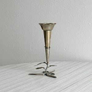 Puede incluir: Un jarrón de metal plateado con una parte superior acampanada y festoneada y un cuerpo delgado en forma de trompeta. El jarrón está sostenido por una base decorativa con hojas y tallos estilizados, creando un diseño elegante de inspiración floral.