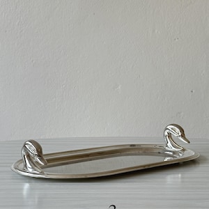 Könnte beinhalten: Ein ovales, silberfarbenes Metalltablett mit zwei geformten Entenkopfgriffen an den gegenüberliegenden Enden. Das Tablett hat eine glatte, reflektierende Oberfläche und einen erhöhten Rand, wobei die Zahl 3 in Schwarz unter dem Objekt abgedruckt ist.