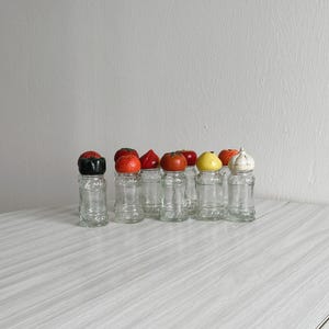 May include: A set of eight clear glass spice jars with colorful vegetable-shaped lids. The lids are red, orange, yellow, and white. The jars are arranged in a row on a light wood surface.