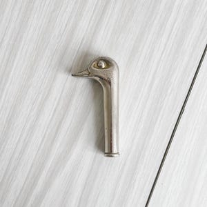May include: A silver-toned bottle opener with a duck head design. The opener features a curved neck and a detailed duck head with an eye and beak. The opener is made of metal and has a vintage or antique appearance.
