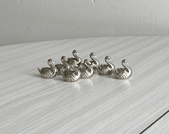 May include: A collection of six small, silver-toned metal swan figurines. Each swan is depicted in a seated position with detailed wing textures and a curved neck. The figures are arranged in a cluster, showcasing a metallic finish and reflective surface.