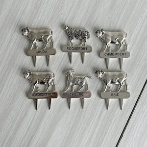 May include: Silver cheese markers with animal designs, including a cow, sheep, and goat. Each marker has a name of a cheese: Gouda, Roquefort, Camembert, Emmental, Chevre, and Brie. The markers are on a light-colored surface.