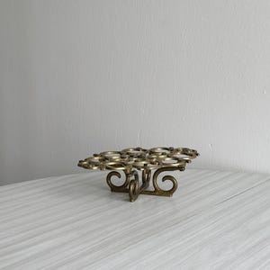 Puede incluir: Un soporte vintage de metal color latón para cupcakes o magdalenas. El soporte presenta un diseño decorativo calado con múltiples soportes circulares. La base tiene un diseño en espiral. El soporte está sobre una superficie clara.