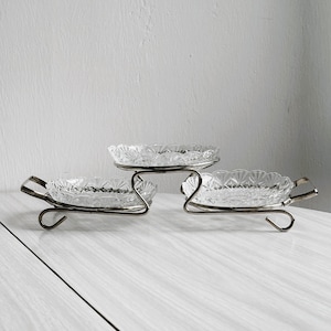 May include: A three-tiered serving dish set. The set features three clear, oval glass dishes with a diamond pattern. The dishes are supported by silver-coloured metal stands with curved handles. Ideal for serving appetizers or desserts.
