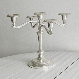 Könnte beinhalten: Ein silberner Kerzenleuchter mit fünf Kerzenhaltern. Der Kerzenleuchter hat einen dekorativen Sockel und einen zentralen Stiel, der sich verzweigt, um die Kerzenbecher zu halten. Die Kerzenbecher haben ein dekoratives, seilartiges Design an den Rändern.