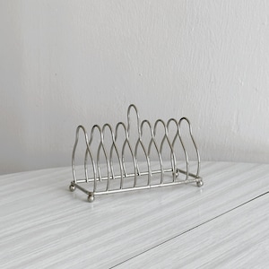 Puede incluir: Un tostador de metal plateado con asa y ocho ranuras. El soporte tiene un diseño sencillo y elegante, perfecto para sujetar rebanadas de pan tostado. El soporte está hecho de alambres de metal finos y curvados.