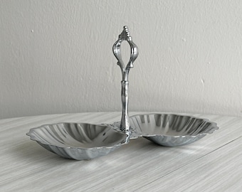 Bandeja metálica ligera de doble compartimento, con forma de concha y asa ornamentada, perfecta para servir aperitivos, pequeños obsequios o como un regalo elegante y original.