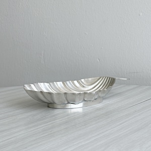 Peut inclure: Un plat en métal argenté en forme de coquillage avec un bord festonné et un rebord lisse et surélevé. Le bol présente un intérieur concave nervuré et repose sur une petite base ovale sur pied, offrant une finition métallique réfléchissante.
