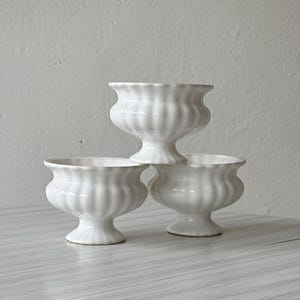 Pode incluir: Três taças de cerâmica branca com um design canelado e estriado e uma base de pedestal. As taças apresentam uma borda larga e curva e um acabamento brilhante. Estão dispostas numa pilha com uma taça sobre as outras duas, mostrando a forma.