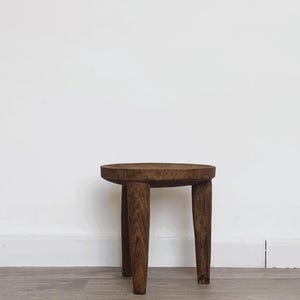 May include: A small, round wooden stool with three tapered legs. The stool is made of dark brown wood and has a simple, rustic design. The top is a flat circle, and the legs are evenly spaced.
