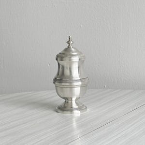 Op de afbeelding: Een zilverkleurige metalen pot met deksel en een gladde, gepolijste afwerking. De houder heeft een ronde voet, een taps toelopend middengedeelte en een afneembaar deksel met een kleine, decoratieve knop. Het deksel en de voet zijn voorzien van fijne, verticale ribbels.