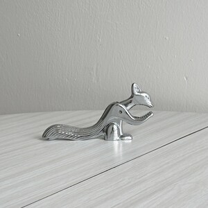 May include: A polished silver squirrel-shaped nutcracker. The nutcracker features a detailed design with a long tail and a squirrel head with open jaws. It is placed on a light-colored wooden surface.