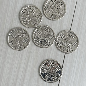 May include: A set of six circular silver-toned metal coasters, each featuring a raised, embossed design of grape clusters and leaves. The coasters have a polished, reflective finish and a scalloped edge, showcasing a consistent botanical motif.