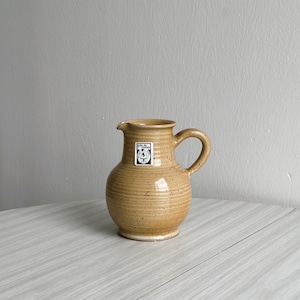 Puede incluir: Una jarra de cerámica de color beige moteada con asa y una pequeña etiqueta en blanco y negro que dice "Porzellan Manufaktur"