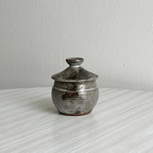 Peut inclure: Un petit pot en céramique fait main, de forme ronde, accompagné de son couvercle assorti. L'objet présente une glaçure terreuse et tachetée dans des tons de gris, de brun et de charbon, avec une finition lisse et brillante.