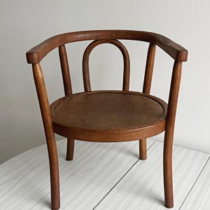 Puede incluir: Silla de madera antigua, pequeña, con respaldo y asiento redondeados. La silla tiene un acabado marrón oscuro y un diseño clásico, con reposabrazos y patas curvadas. El asiento es circular y parece ser de madera maciza.