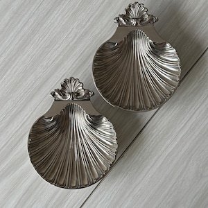 Peut inclure: Deux coupelles en métal argenté en forme de coquille Saint-Jacques. Chaque coupelle présente une surface polie et réfléchissante avec des cannelures profondes et rayonnantes, ainsi qu'une crête ornementale en éventail sur le bord supérieur.