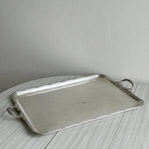 May include: A rectangular silver-toned serving tray with rounded corners and two ornate handles on the short sides. The tray features a raised border with a subtle embossed pattern along the edges and a smooth, reflective central surface area.