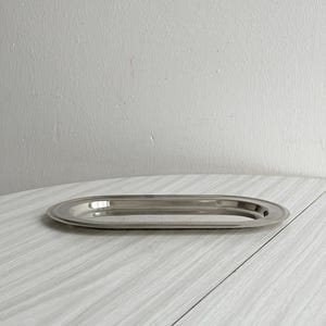 Può includere: Un vassoio ovale in acciaio inossidabile lucido. Il vassoio ha un bordo rialzato e una superficie riflettente. Si trova su una superficie a grana di legno grigio chiaro. Il vassoio è adatto per servire cibo o esporre oggetti.